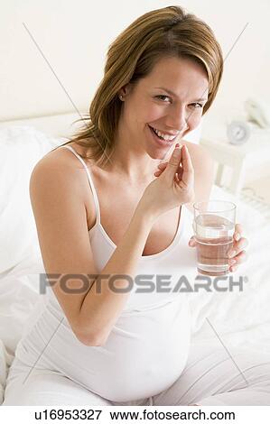Stock Photo - Pregnant woman in bedroom taking medicine holding water smiling. Fotosearch
