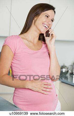 Stock Photo - Pregnant woman in kitchen talking on cellular phone and smiling. Fotosearch