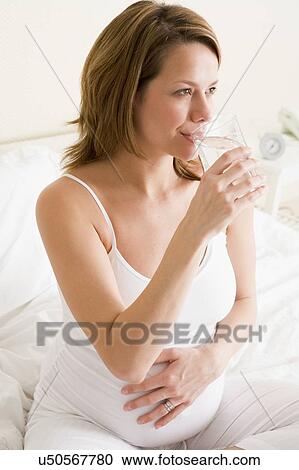 Stock Image - Pregnant woman sitting in bedroom with glass of water smiling. Fotosearch