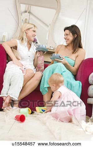 Stock Photo - Two mothers in living room with babies and coffee smiling. Fotosearch
