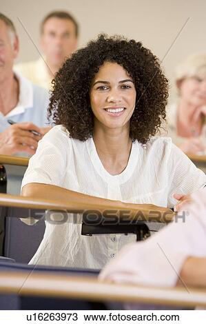 Stock Image - Woman sitting in adult classroom with students in background (selective focus). Fotosearch