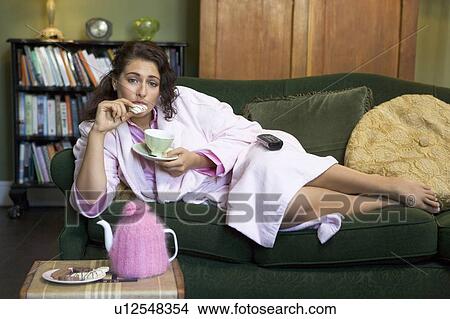 Young woman lying on sofa at home eating cookies and drinking tea View Large Photo Image Picture - Young woman lying on sofa at home eating cookies and drinking tea. Fotosearch
