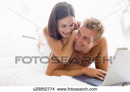 Picture - Couple lying in bed with laptop smiling. Fotosearch