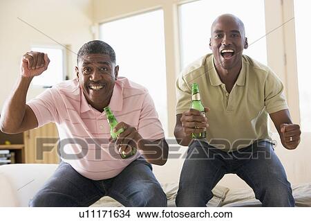 Picture - Two men in living room with beer bottles cheering and smiling. Fotosearch