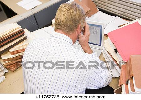 Stock Photo - Businessman in cubicle with laptop and stacks of files. Fotosearch