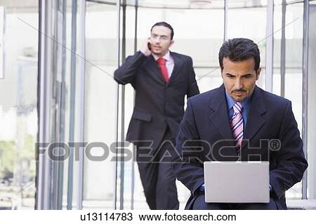Businessman outdoors in front of building using laptop with businessman in background on cellular phone (high key/selective focus) View Large Photo Image Stock Image - Businessman outdoors in front of building using laptop with businessman in background on cellular phone (high key/selective focus). Fotosearch