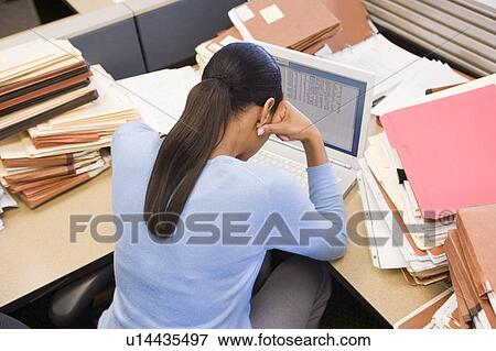 Stock Photo - Businesswoman in cubicle with laptop and stacks of files. Fotosearch