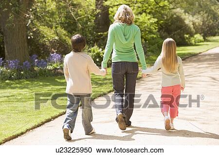 Stock Image - Mother and two young children walking on path holding hands smiling. Fotosearch