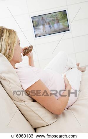 Stock Image - Pregnant woman watching television and eating chocolate smiling. Fotosearch