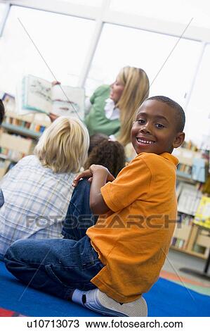 Stock Image - Student in class looking at camera with teacher reading in background (selective focus). Fotosearch