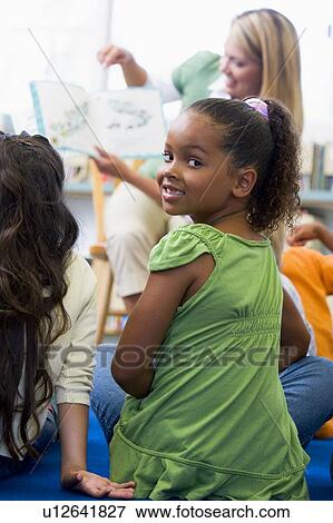 Stock Photo - Student in class looking at camera with teacher reading in background (selective focus). Fotosearch