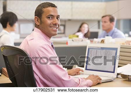 Stock Image - Businessman in cubicle with laptop smiling. Fotosearch