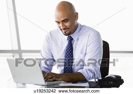 Stock Image - Businessman in office with laptop smiling (high key/selective focus). Fotosearch