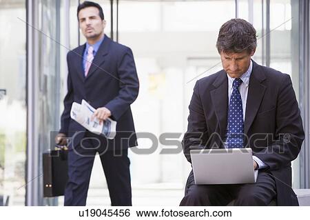 Businessman outdoors in front of building using laptop with businessman in background carrying newspaper (high key/selective focus) View Large Photo Image Stock Photograph - Businessman outdoors in front of building using laptop with businessman in background carrying newspaper (high key/selective focus). Fotosearch