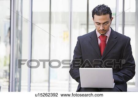 Stock Image - Businessman sitting outdoors by building with a laptop (high key/selective focus). Fotosearch