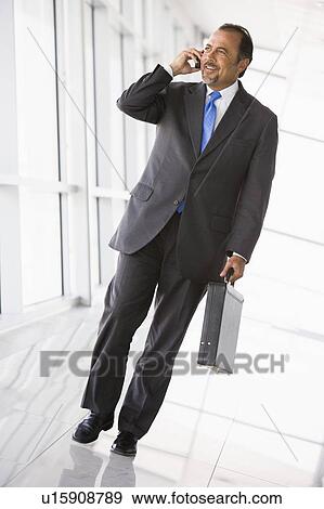 Businessman walking in corridor on cellular phone smiling (high key/selective focus) View Large Photo Image Stock Photo - Businessman walking in corridor on cellular phone smiling (high key/selective focus). Fotosearch