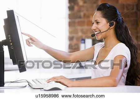 Stock Photograph - Businesswoman in office wearing headset and pointing at computer monitor. Fotosearch