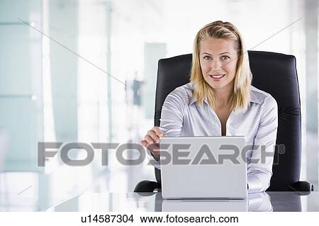 Picture - Businesswoman sitting in office with laptop smiling. Fotosearch
