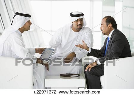 Stock Image - Three businessmen indoors with a laptop talking (high key/selective focus). Fotosearch