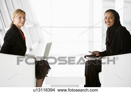Two businesswomen indoors with a laptop smiling (high key/selective focus) View Large Photo Image Picture - Two businesswomen indoors with a laptop smiling (high key/selective focus). Fotosearch