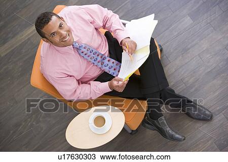 Stock Image - Businessman sitting indoors with coffee and folder smiling. Fotosearch