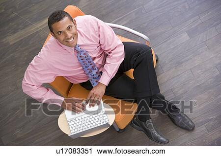 Businessman sitting indoors with laptop smiling View Large Photo Image Stock Image - Businessman sitting indoors with laptop smiling. Fotosearch