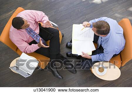Stock Image - Two businessmen sitting indoors with coffee laptop and folder. Fotosearch