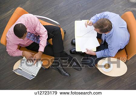 Stock Photo - Two businessmen sitting indoors with coffee laptop and folder. Fotosearch