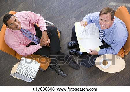 Stock Photo - Two businessmen sitting indoors with coffee laptop and folder smiling. Fotosearch