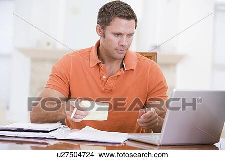 Picture - Man in dining room with laptop holding paperwork. Fotosearch