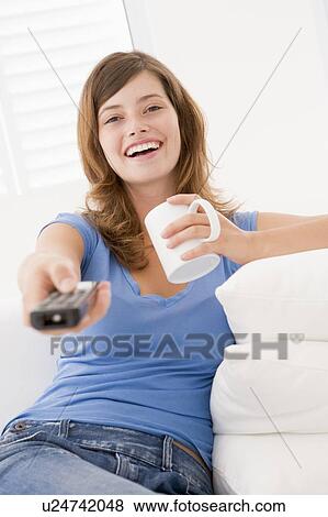 Stock Photo - Woman in living room with remote control and coffee smiling. Fotosearch