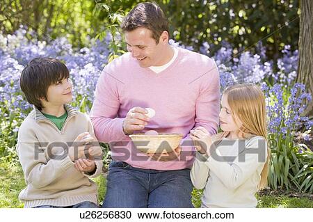 Stock Image - Father and two young children on Easter looking for eggs outdoors smiling. Fotosearch
