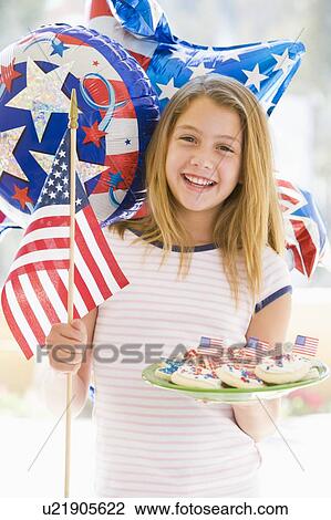 Stock Image - Young girl outdoors on fourth of July with flag and cookies smiling. Fotosearch