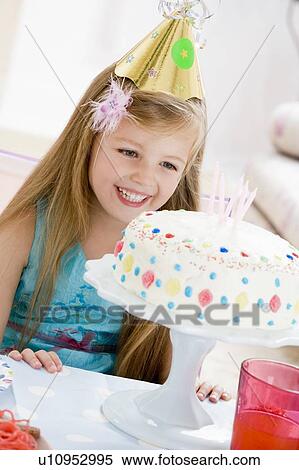 Stock Photography - Young girl wearing party hat looking at birthday cake smiling. Fotosearch