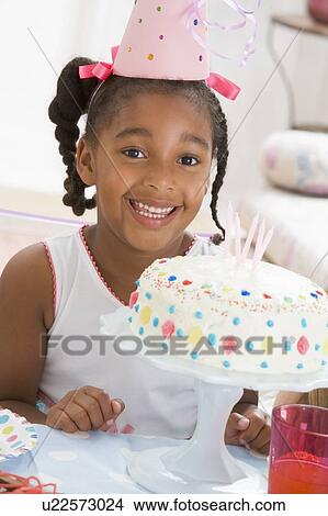 Picture - Young girl wearing party hat with cake in front of her smiling. Fotosearch