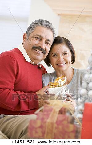 Stock Image - Middle Aged Couple Sitting On Sofa Holding Christmas Present. Fotosearch