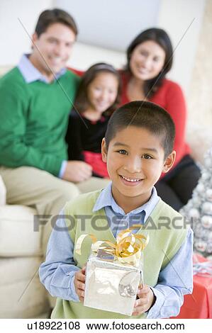 Stock Photo - Young Boy Standing Holding Christmas Present, With His Parents And Sister In The Background. Fotosearch