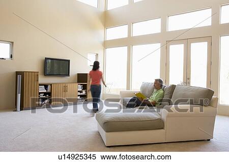Stock Photography - Man reading book in living room with woman walking by. Fotosearch