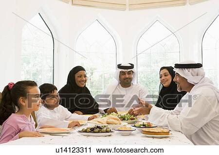 Stock Image - Family sitting at dinner table smiling (high key). Fotosearch