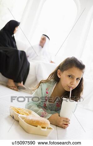 Stock Image - Young girl with fast food in living room with parents in background (high key/selective focus). Fotosearch