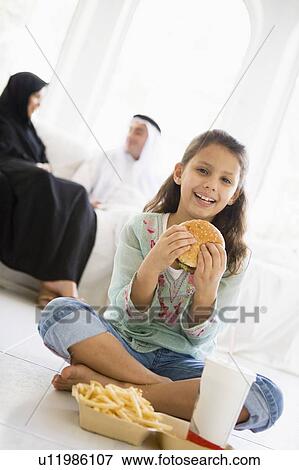 Stock Photo - Young girl with fast food in living room smiling with parents in background (high key/selective focus). Fotosearch