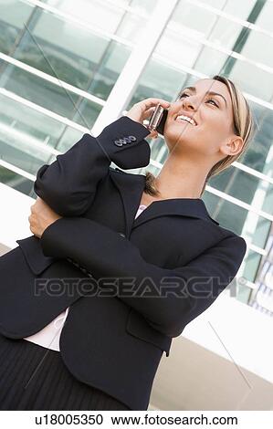 Stock Image - Businesswoman standing outdoors using cellular phone and smiling. Fotosearch