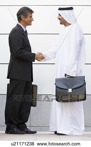 Stock Photo - Two businessmen standing outdoors with briefcases shaking hands and smiling. Fotosearch