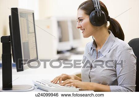 Stock Photograph - Woman wearing headphones in computer room typing and smiling. Fotosearch