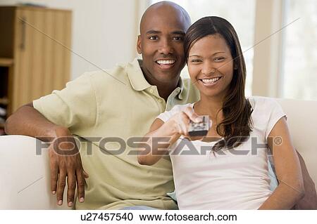 Stock Image - Couple in living room with remote control smiling. Fotosearch