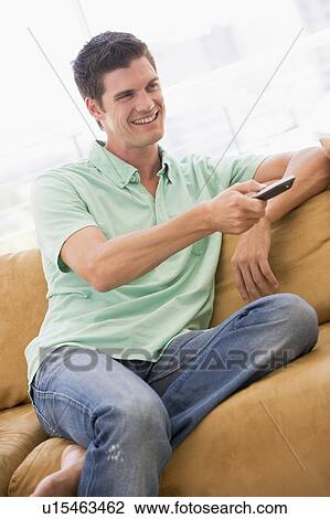 Stock Image - Man in living room with remote control smiling. Fotosearch