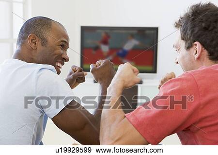 Stock Photo - Two men in living room watching television and cheering. Fotosearch