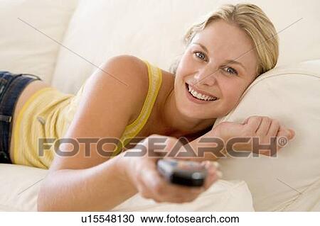 Stock Image - Woman in living room holding remote control smiling. Fotosearch