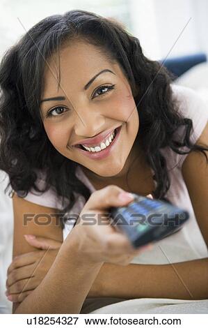 Stock Photo - Woman in living room holding remote control smiling (high key/selective focus). Fotosearch