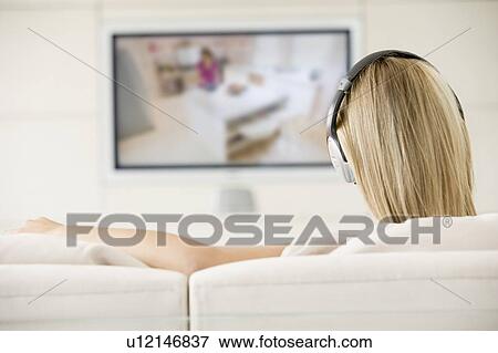 Stock Photo - Woman in living room watching television and wearing headphones. Fotosearch